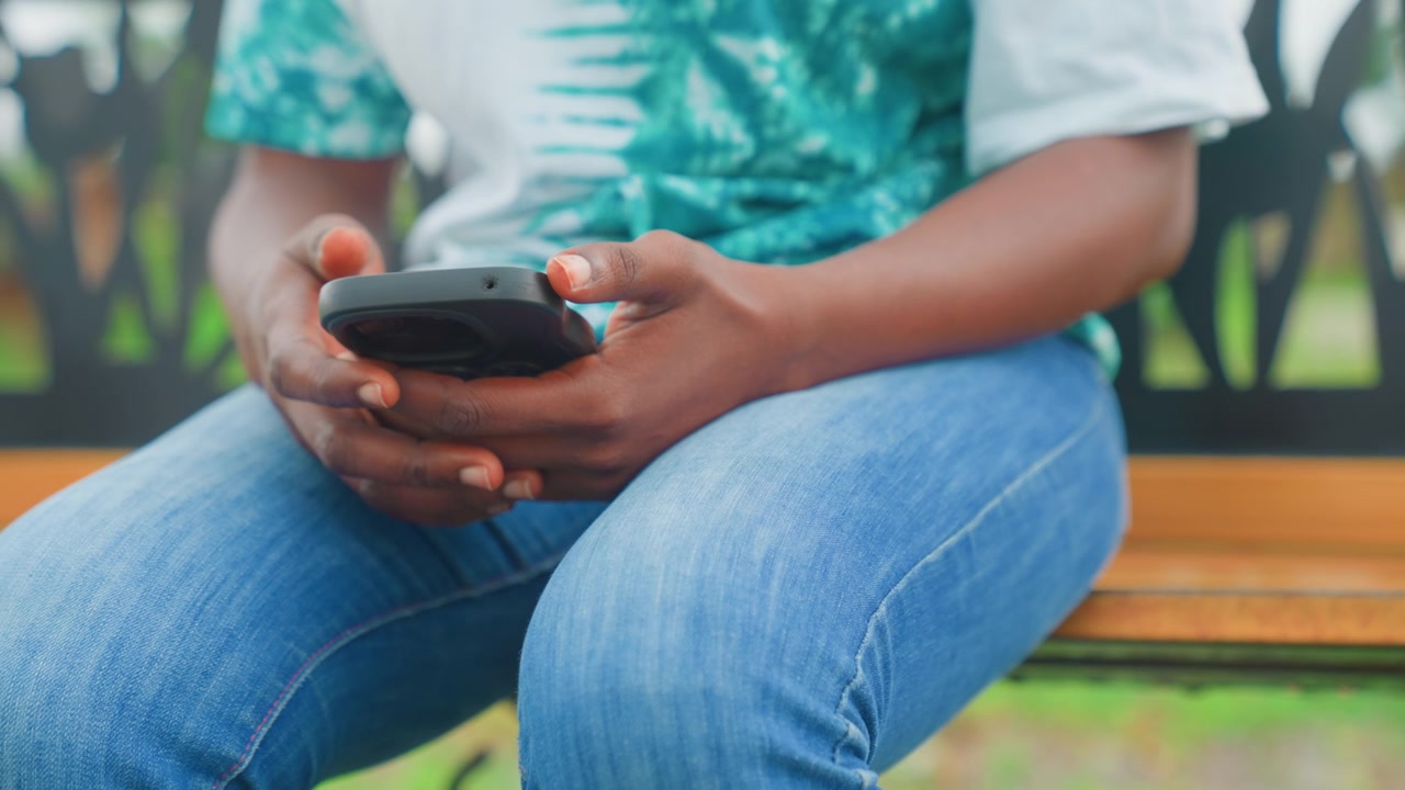 Black mother on bench checking phone, closeup of hands and smartphone, denim jeans and patterned top, natural light and soft bokeh, waiting to pick up child, texting and reading notifications