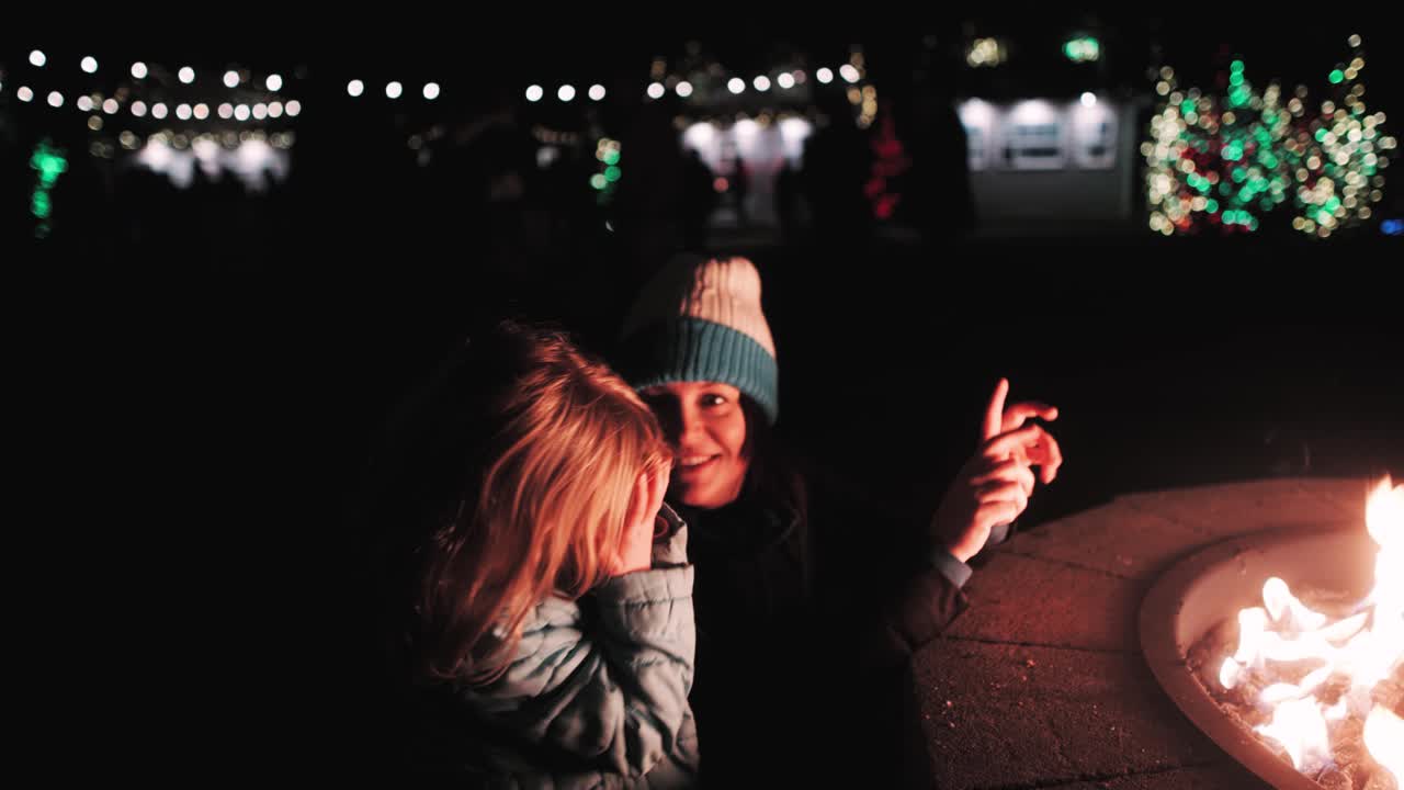 Mother and daughter, three-year old, warming their fingers at a firepit with Christmas lights in the background