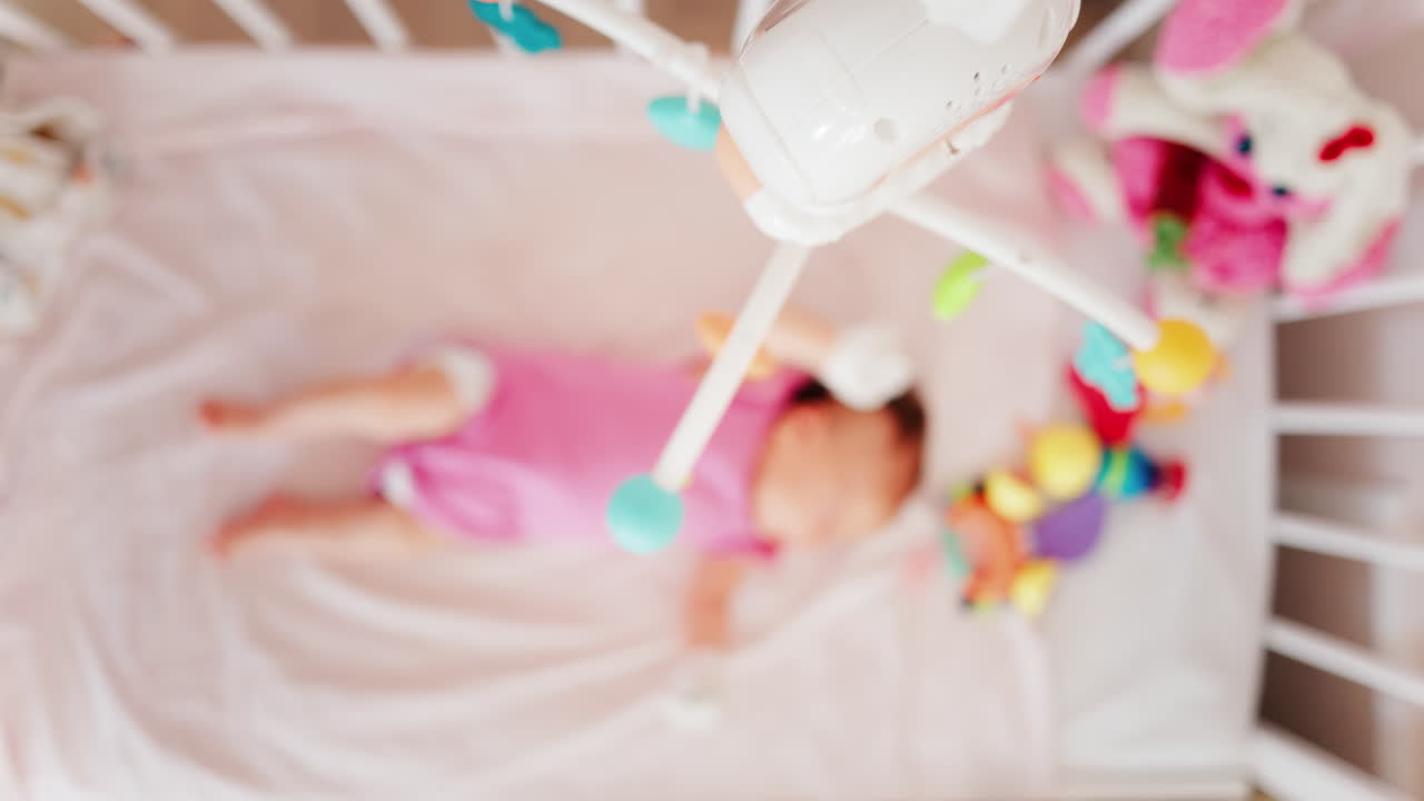 Bambino sdraiato su un letto con un vestito rosa, che guarda un colorato gioco mobile appeso