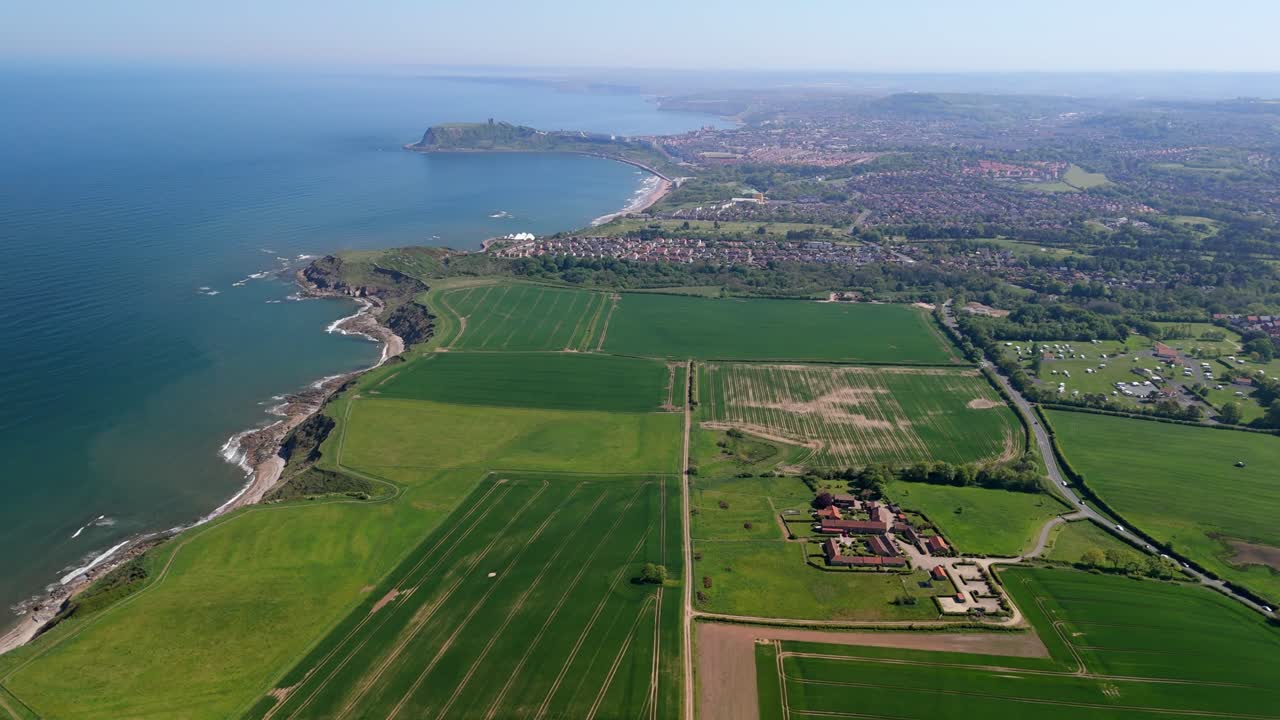 A high-angle drone shot captures a slow approach over farmland towards the entire coastline of Scarborough and the Castle