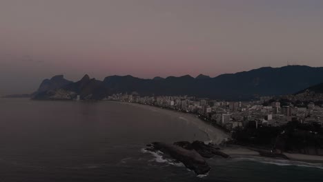 Aerial-approach-and-camera-tilt-down-showing-the-Arpoador,-harpooner,-cliff-off-of-the-coast-of-Rio-de-Janeiro-at-sunrise-with-the-Ipanema-neighbourhood-in-the-background