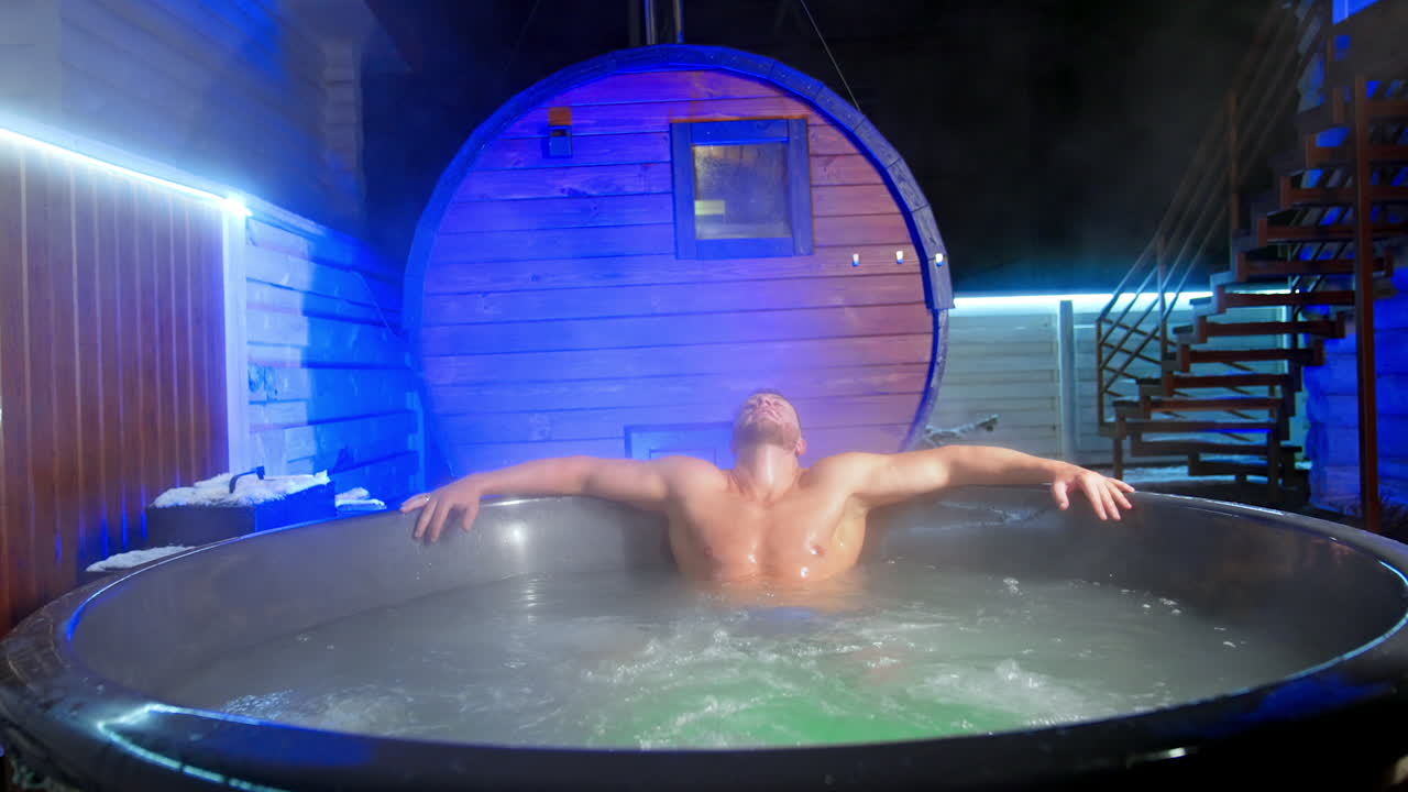 Caucasian man rubs his face sitting in hot tub. Man leans back relaxing in Jacuzzi outdoors in winter.