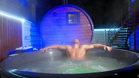 caucasian man rubs his face sitting in hot tub. man leans back relaxing in jacuzzi outdoors in winter.