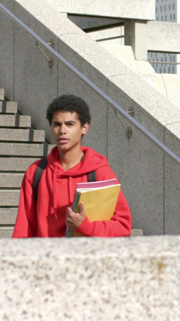 Video verticale: Studente che inizia a scendere, con delle cartelle in mano e una felpa rossa con cappuccio, guardando a destra sulle scale della piazza