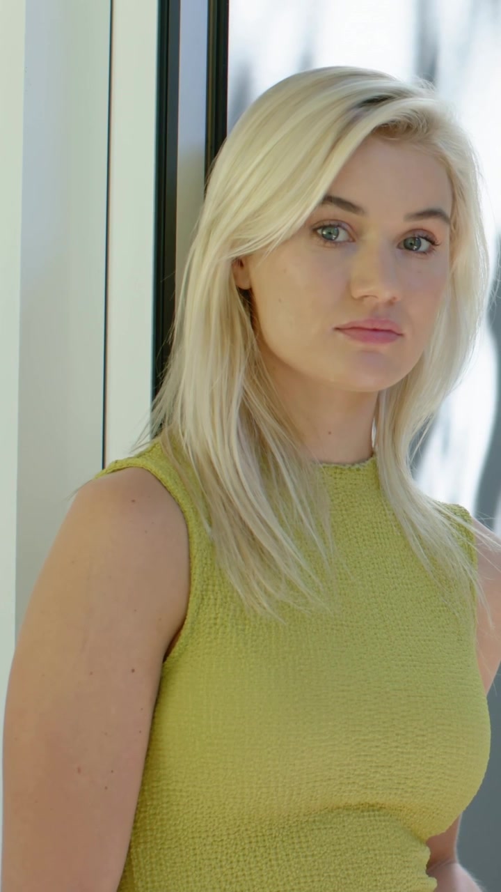 Vídeo vertical: Mulher com blusa verde, iluminada por luz suave, virando-se para a câmera e sorrindo junto a um painel de vidro.