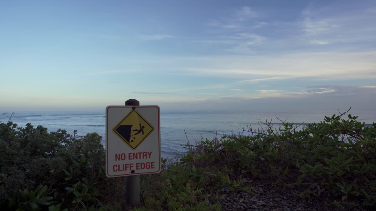 Cartello di divieto d'accesso, dirupo a strapiombo lungo la Great Ocean Road, costa tranquilla e luce soffusa della sera mentre le onde si infrangono sulla riva