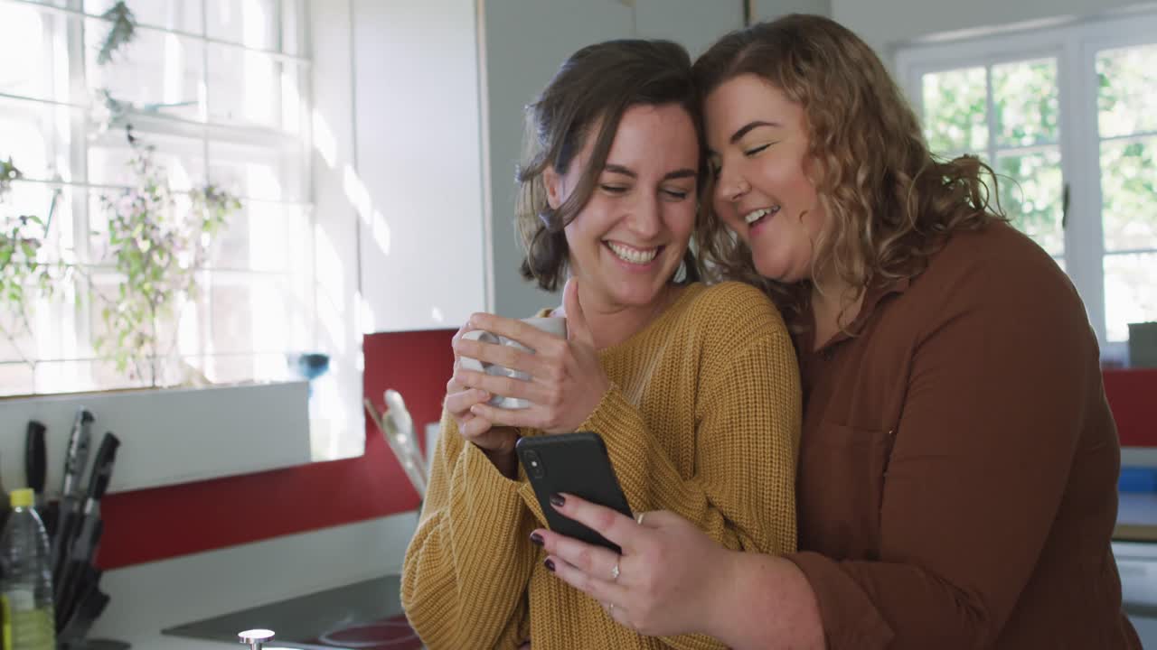 feliz pareja de lesbianas caucásicas abrazándose y usando un teléfono inteligente en la cocina