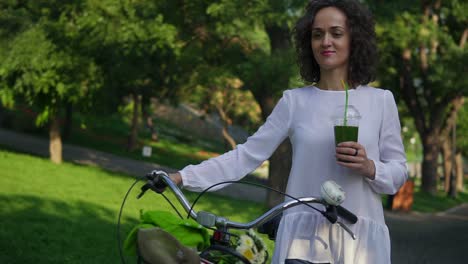 Beautiful-woman-in-a-white-shirt-and-blue-jeans-walking-in-the-city-park-holding-her-city-bicycle-handlebar-with-flowers-in