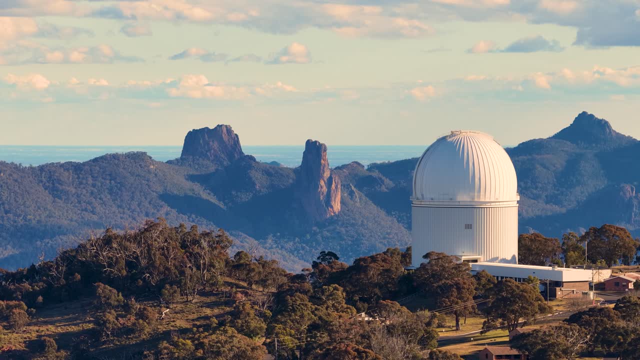 Aerial drone footage pans across Siding Spring Observatory domes and rugged Warrumbungle mountain landscape in warm sunset light, with clear skies and dramatic terrain