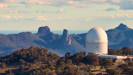 aerial drone footage pans across siding spring observatory domes and rugged warrumbungle mountain landscape in warm sunset light, with clear skies and dramatic terrain