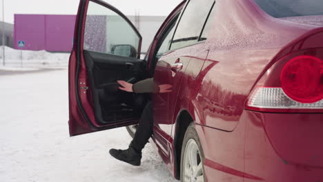civil engineer wearing winter gear exits red car parked on snowy lot and walks away as light snow falls, with urban signposts and industrial buildings visible in soft distant background