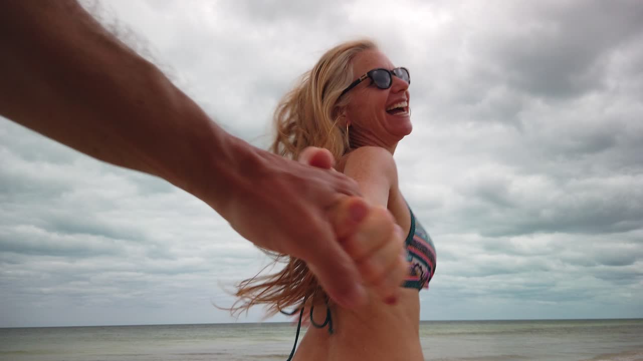 mujer madura correr bikini y gafas de sol corriendo en cámara lenta en sígueme tirando de su marido en la playa