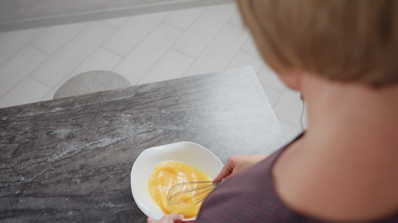 vista trasera de la dama con el cabello dorado usando un vestido a cuadros agitando el huevo en el cuenco, ella levanta el batidor ligeramente antes de continuar mezclando, con una encimera lisa y iluminación suave