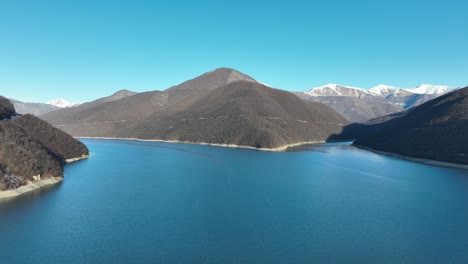 A vast reservoir nestled in snowy mountain valleys, basking in soft sunlight. The vibrant blue water contrasts with the rugged, tree-lined slopes