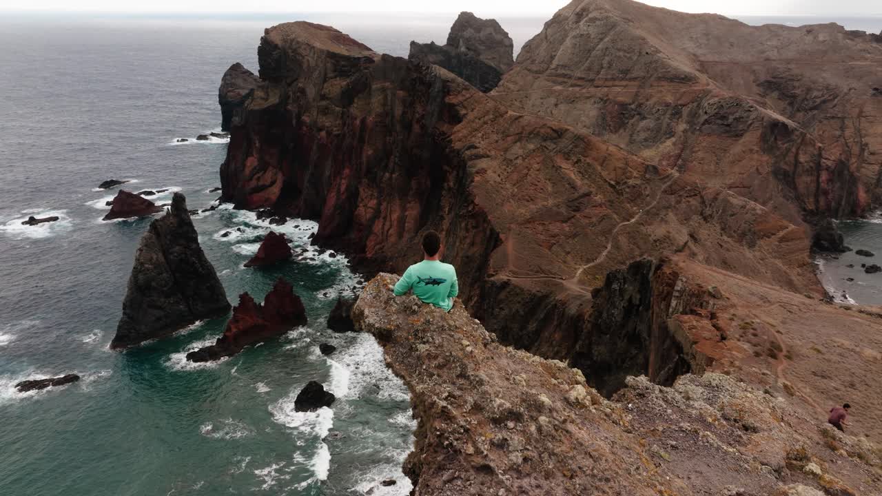 Aerial orbit around a man sitting on a cliff, capturing the dramatic coastline and ocean view of Ponta de São Lourenço.