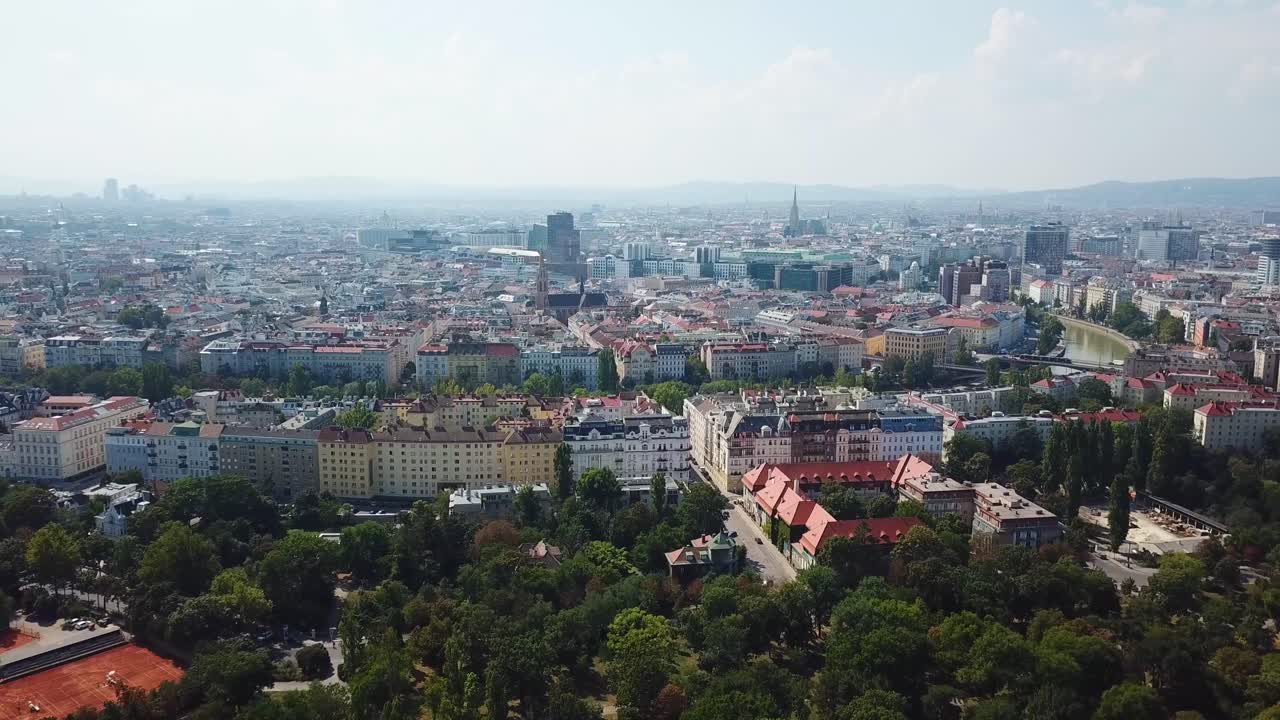 Aerial approaching shot of colorful trees of park in suburbia of Vienna City. Row of colored apartment houses and homes with river view. Sunny day in capital of Austria. Skyline downtown in background