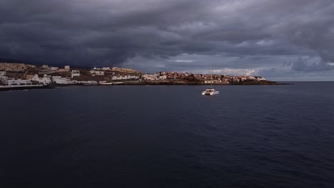 Picturesque-aerial,-Town-with-houses-and-windmills-at-coast-beach-of-island-tenerife,-sail-boat-in-water-near-coast