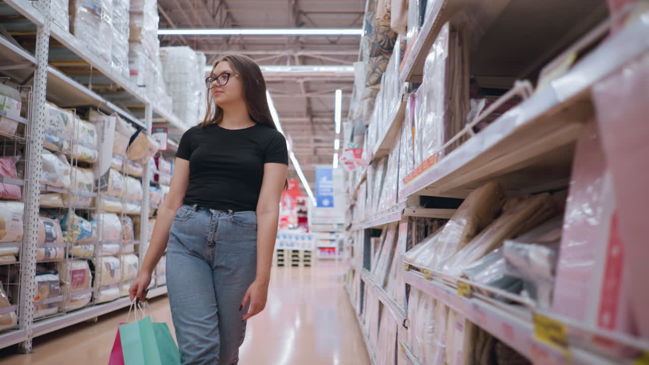 mujer joven de camiseta negra caminando cerca de la estantería de la tienda, observando los productos mientras lleva bolsas de compras, fondo borroso con otro comprador empujando el carrito
