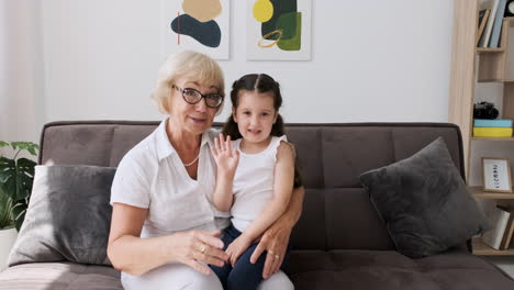 Abuela-Feliz-Con-Su-Linda-Nieta-Sentada-De-Rodillas-En-El-Sofá-Saludando-Y-Hablando-Por-Videollamada