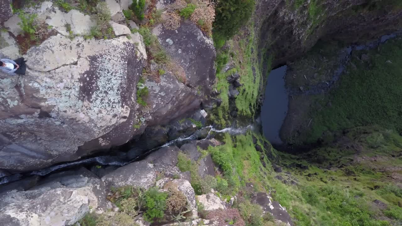 madeira, portugal - hombre sentado en la roca en la parte superior del acantilado mirando el agua que fluye hacia la cascada - toma aérea de drones