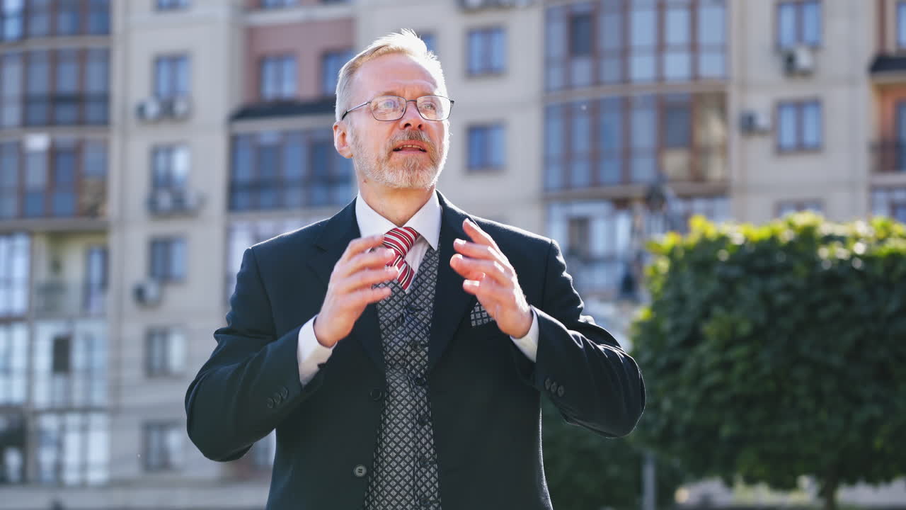 Portrait senior businessman in eyeglasses on the building background in the city. Serious bearded man with grey hair in suit telling about his business outdoors.