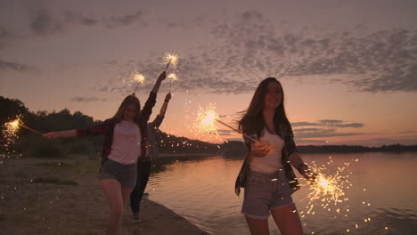 Cheerful-male-and-female-friends-are-running-along-the-beach-at-sunset-holding-sparkling-fireworks-and-runaway-lights-in-slow-motion.-Dancing-and-sunset-party-on-the-beach