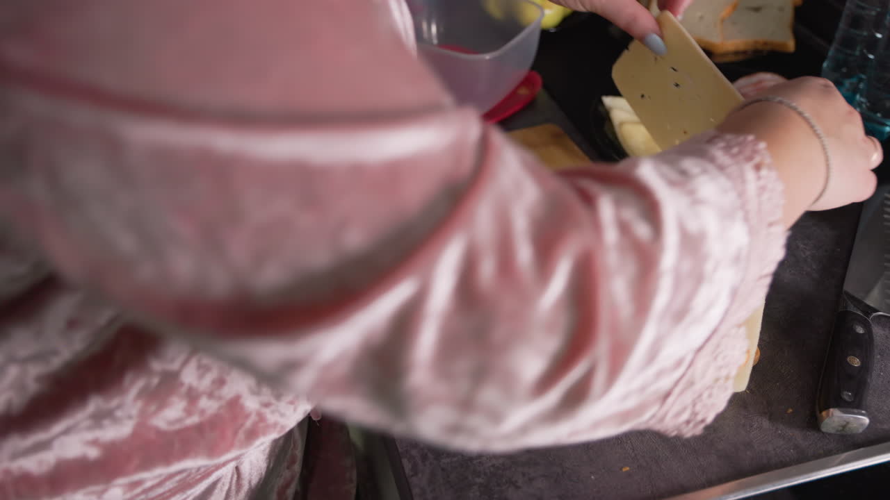 Caregiver in pink robe prepares sandwich on wooden cutting board, layering slices of cheese and bread while working over kitchen countertop, hands visible in frame beside black knife and dark surface