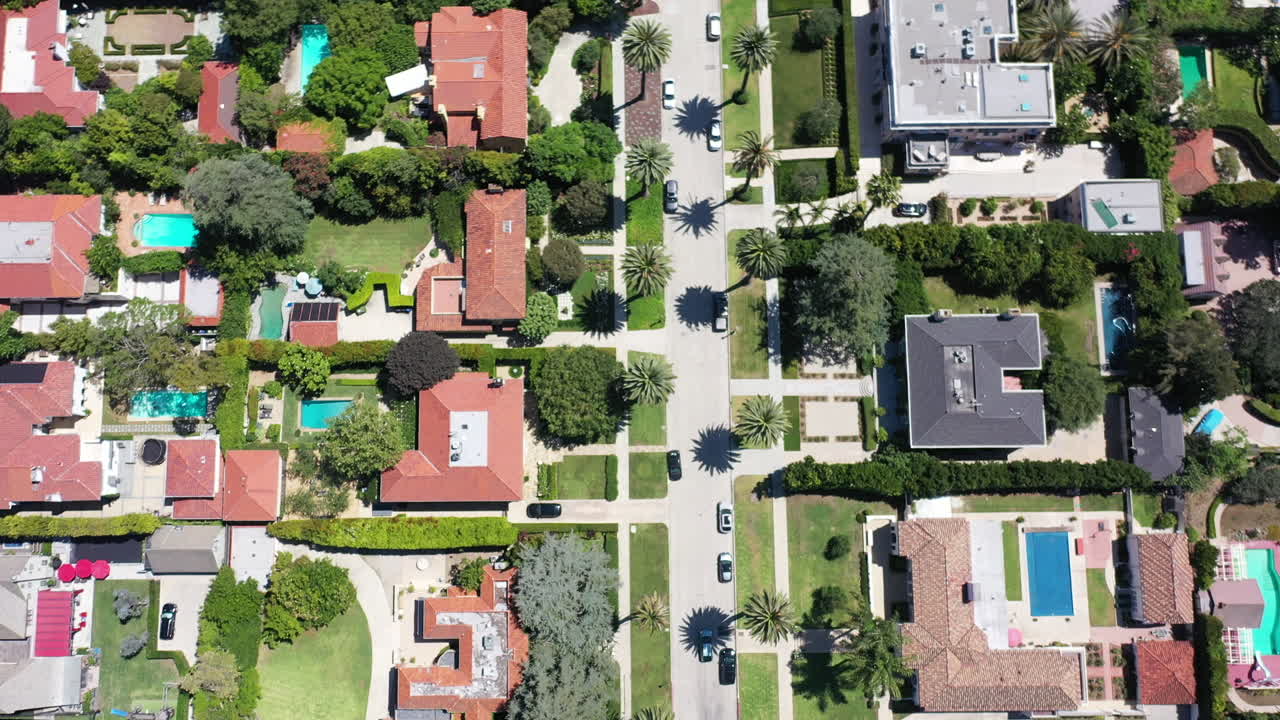películas de drones desde lo alto, siguiendo lentamente a un automóvil blanco que conduce por una calle bordeada de palmeras, rodeado de casas en un hermoso vecindario de west hollywood, california