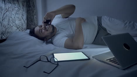 an indian young man lying on the bed and talking on mobile phone after using laptop for long time at night