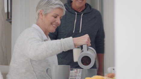 happy mature couple eating breakfast together husband looking for milk in fridge sitting with wife at table sharing morning meal in kitchen day in the life 4k footage