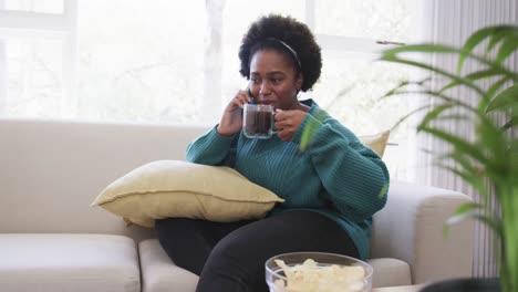 Fröhliche-Afroamerikanische-Frau-Entspannt-Sich-Auf-Der-Couch,-Spricht-Auf-Dem-Smartphone-Und-Trinkt-Tee,-Zeitlupe