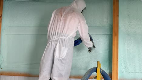 worker in respirator and tyvek suit sprays second closed cell foam insulation on the house wrap and studs of newly constructed exterior wall