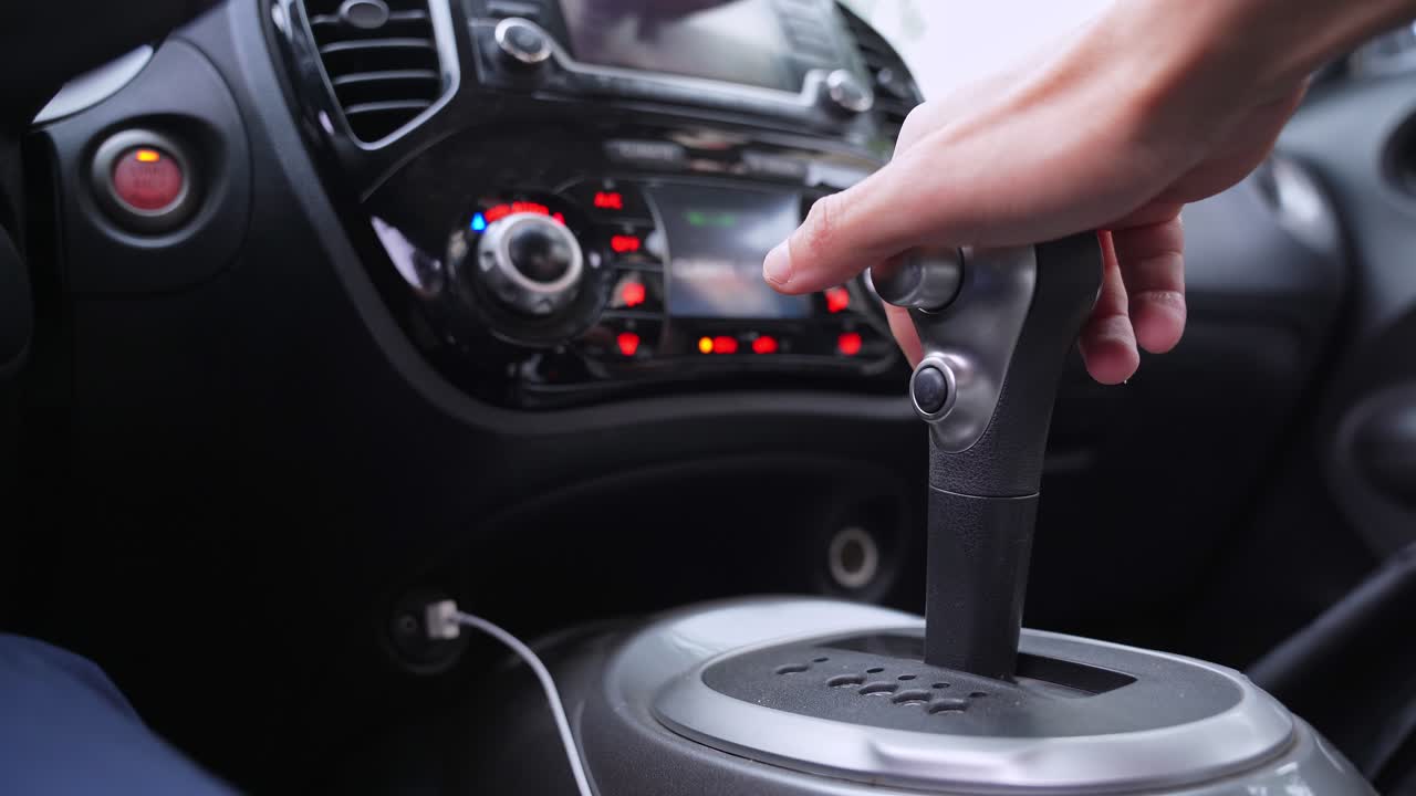 最新の車両の車内で、運転手がオートマチックのギアレバーを操作している様子。自然光が差し込む中、気候制御機能付きのダッシュボードがぼやけた背景に見える。