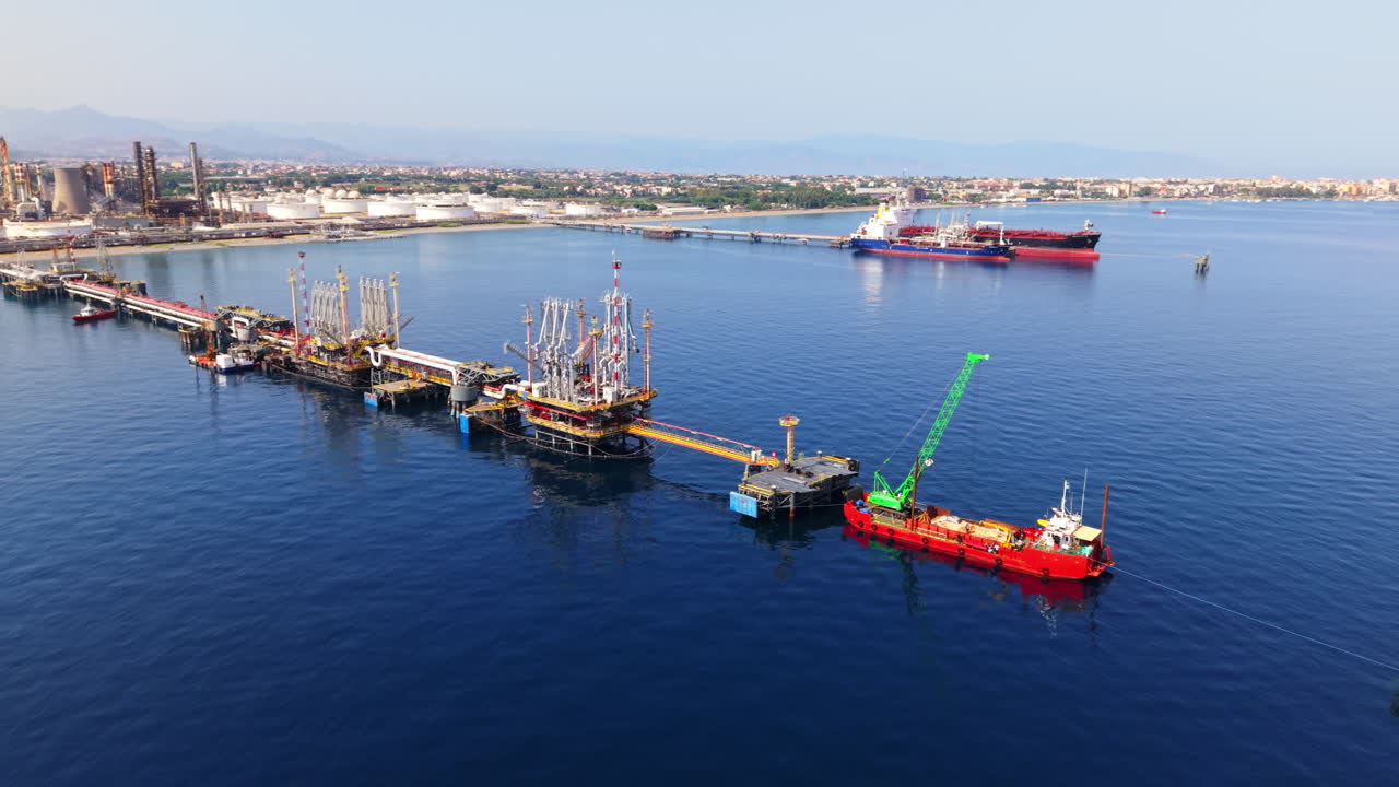 Drone shot approaching offshore oil terminal with ships, refinery tanks, and Sicilian coastline