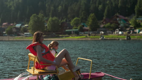 mother and daughter pedale catamaran moving near shore. river with lush green vegetation and tourist lodges side view. woman and child rest enjoying movement on water slow motion