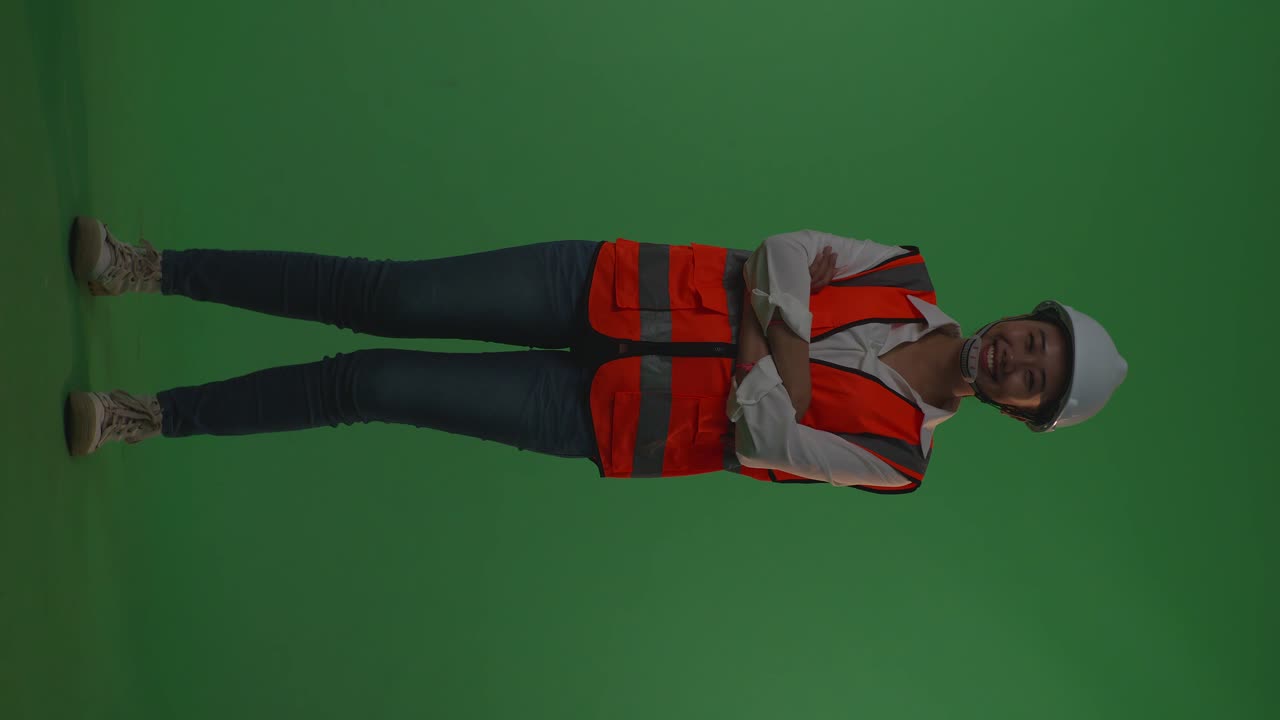 Full Body Of Asian Female Engineer With Safety Helmet Crossing Her Arms And Smiling To Camera While Standing In The Green Screen Background Studio, During Sunset Or Sunrise Time
