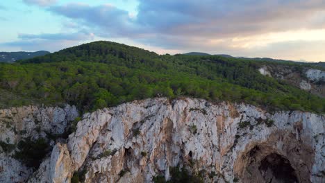 Hikers-rest-on-a-rocky-cliff-among-trees-with-a-view-of-the-green-landscape-during-sunset-in-ibiza