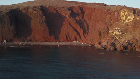 Aerial-flight-to-Paralia-deep-red-beach-cliffs,-Santorini-south-coast