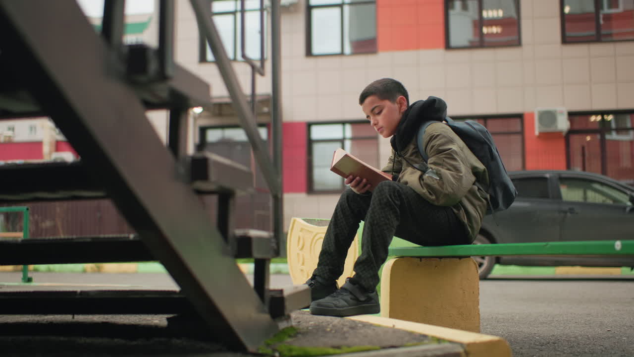 High school student seated on bench with backpack flipping through book while studying outdoors near blurred urban building and glowing bulb on door post and parked car