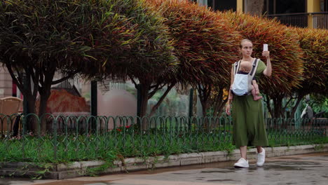 A-young-mother-with-her-baby-in-a-kangaroo-backpack-takes-photos-using-a-mobile-phone-while-traveling.-She-walks-and-occasionally-glances-at-the-phone-screen