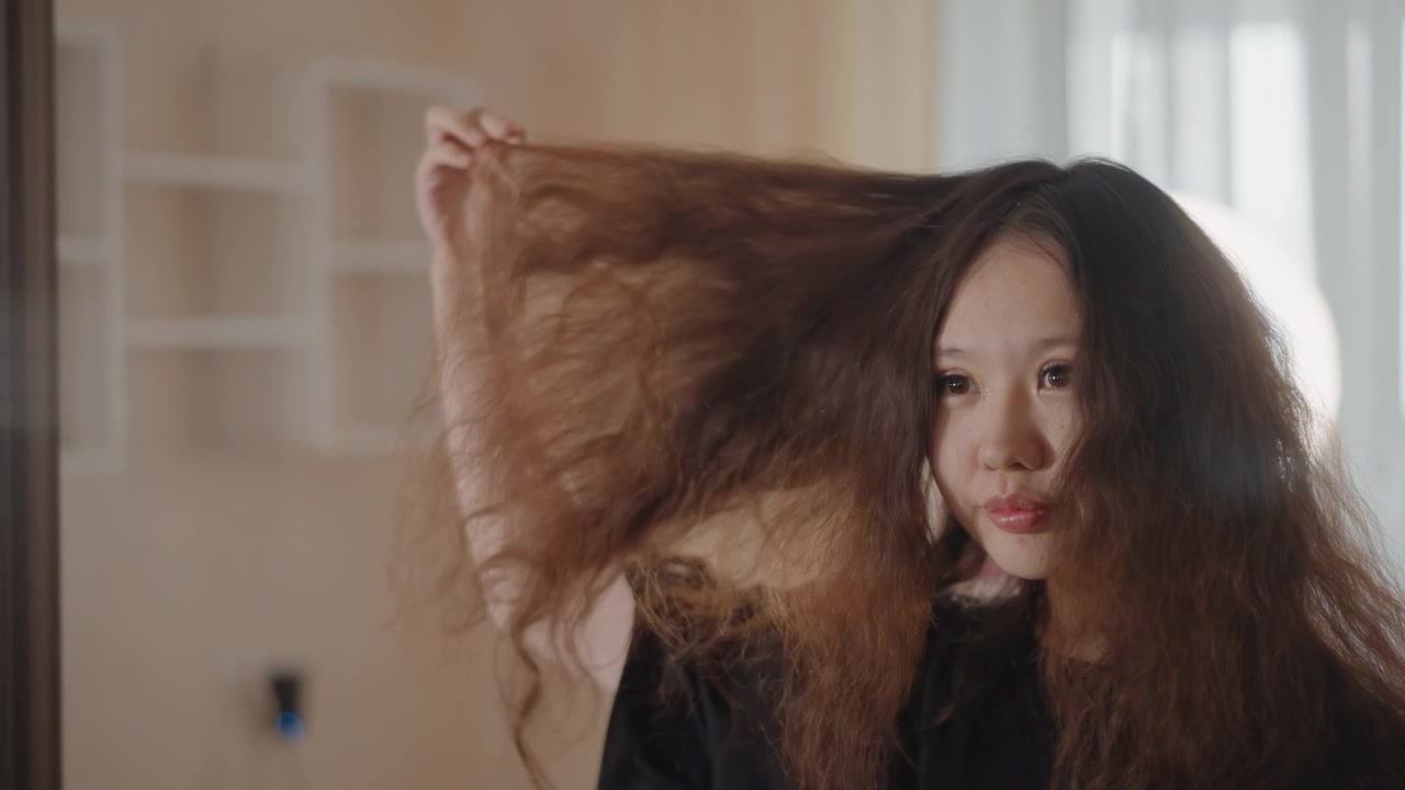 Wanita muda merawat rambut keritingnya yang panjang dan bervolume sambil melihat ke arah cermin, menyisir, mengangkat, menyesuaikan, dan dengan lembut merapikan helai rambut selama rutinitas kecantikan pribadi yang fokus di interior yang remang-remang.
