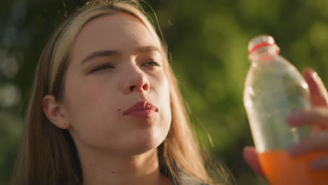 mujer al aire libre bebiendo jugo de naranja de una botella de plástico con contenido y expresión relajada, rodeada de naturaleza y luz solar suave, con vegetación borrosa en el fondo