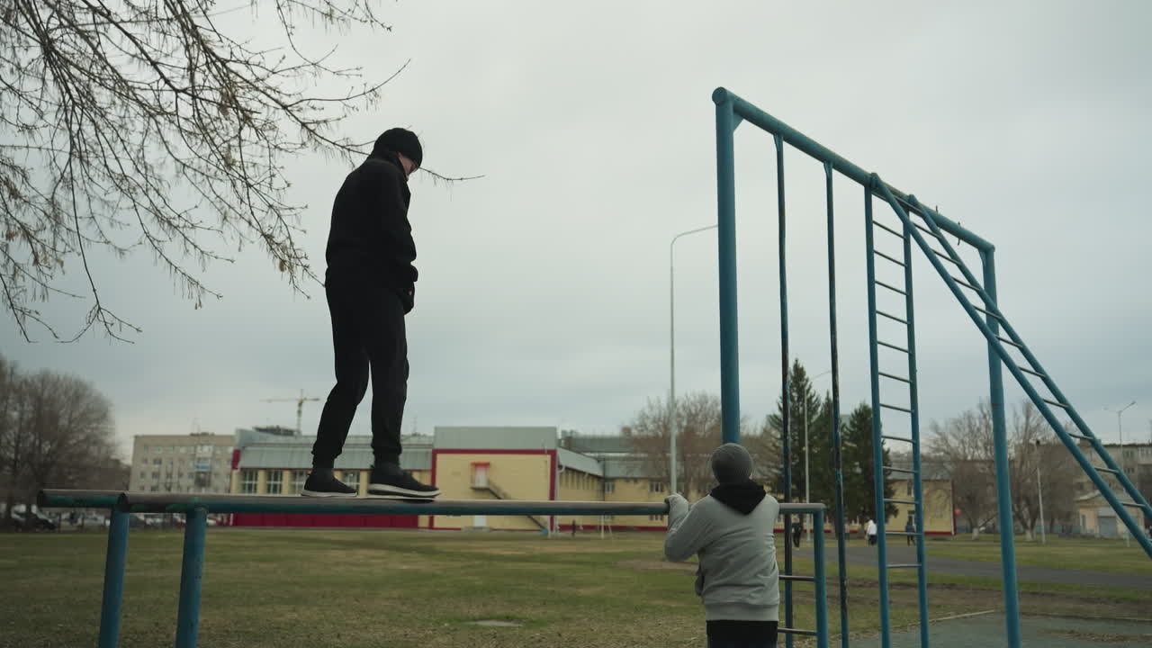 A young boy dressed in a black outfit carefully walks along an iron pole carefully, while another person, dressed in a gray jacket and beanie, rests against the pole, looking up