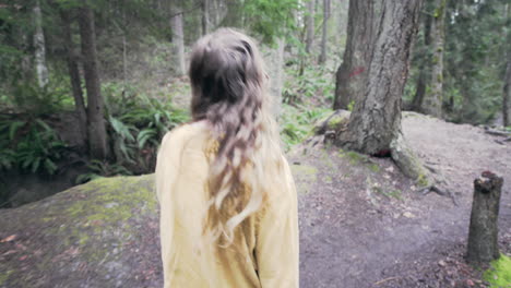 a young woman wearing a bright yellow jacket walks over a small bridge in a forest, tracking shot from behind