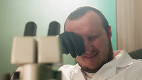 una vista de cerca de un científico con una bata blanca de laboratorio estremecéndose y usando sus manos para limpiar sus ojos, mientras examina muestras a través de un microscopio en un laboratorio