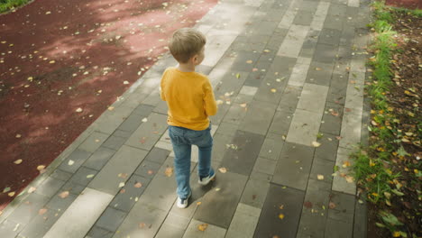rear view of young boy in yellow long-sleeve shirt and jeans walking alone on paved path in peaceful park, glancing around with curiosity as autumn leaves scatter across shaded ground