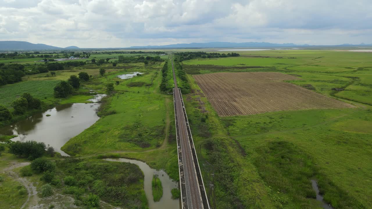 Reverse aerial footage showing a lake on the left, farmlands, dirt roads, picturesque horizon; Elevated railways with a gorgeous landscape and lake, in Saraburi, Thailand.