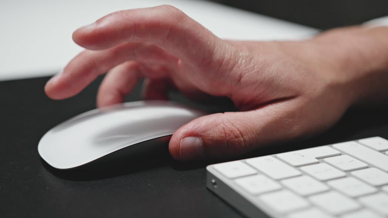 A close-up shot of a person's hand using a modern, white wireless mouse on a black mouse pad. The fingers are actively clicking and scrolling, with a sleek keyboard nearby. Tech and office concepts.