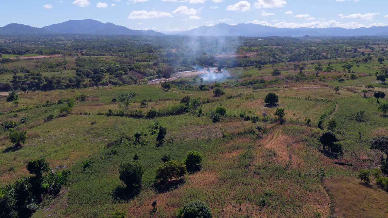 Aerial view of burning land in Haiti under bright sunlight, showing smoke rising over rural fields and hills, sad environmental destruction, deforestation, and ecological impact in countryside