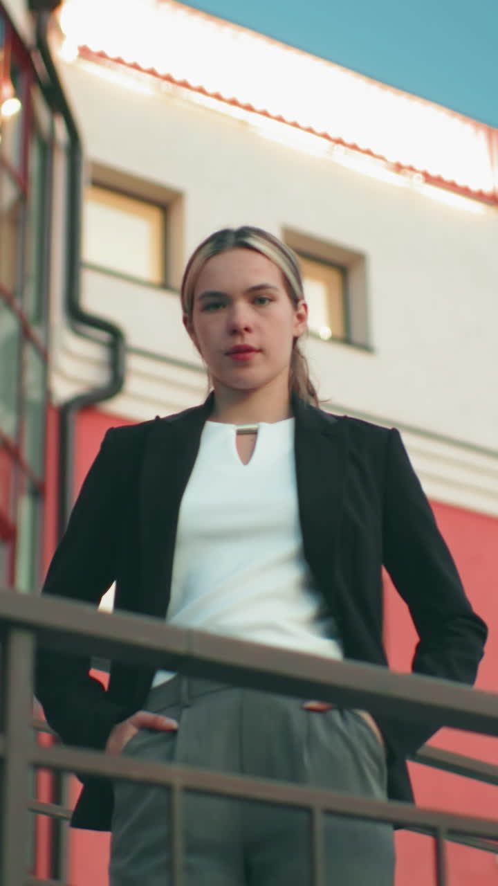 Down top view of woman confidently posing in front of camera with one hand in pocket outside modern building with reflective glass and display light blinking on roof top under clear blue sky